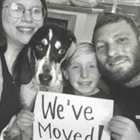 Black and white photo of couple with their child and dog, holding a sign that says "We've Moved"