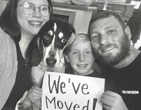 Black and white photo of couple with their child and dog, holding a sign that says "We've Moved"