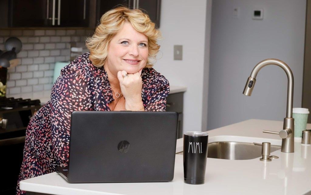 Wendy Wendorf leaning on counter with her hand under her chin smiling, laptop and coffee mug with "MIMI" on it